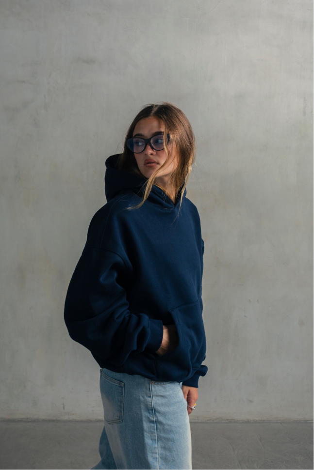 Model wearing a navy blue relaxed-fit hoodie with light blue jeans, shown in a side profile with hands in the kangaroo pocket.