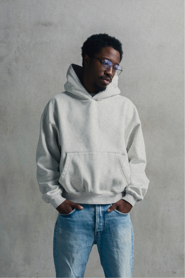 Model wearing a light heather grey relaxed-fit hoodie with light blue jeans, standing with hands in the kangaroo pocket.