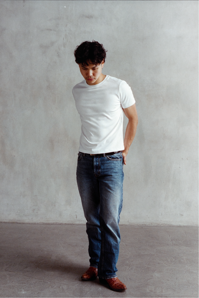 Model wearing a white fitted crew neck T-shirt with blue jeans, looking downward with hands behind the back.