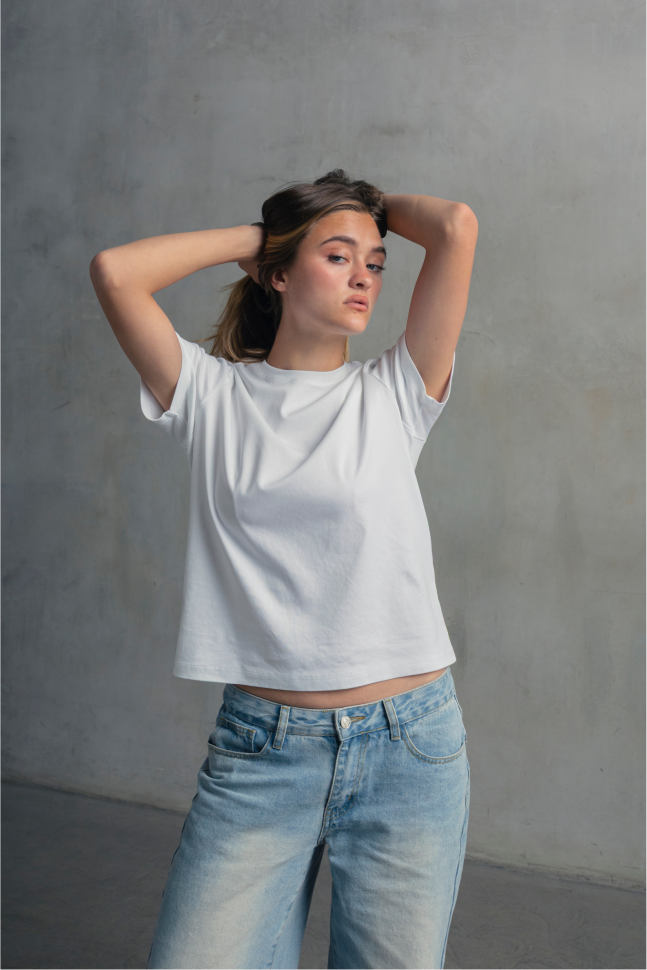 Model wearing a white crew neck T-shirt with light blue jeans, posing with both hands behind her head.