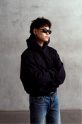 Model wearing a black relaxed-fit hoodie with blue jeans and sunglasses, shown in a three-quarter view with folded arms.