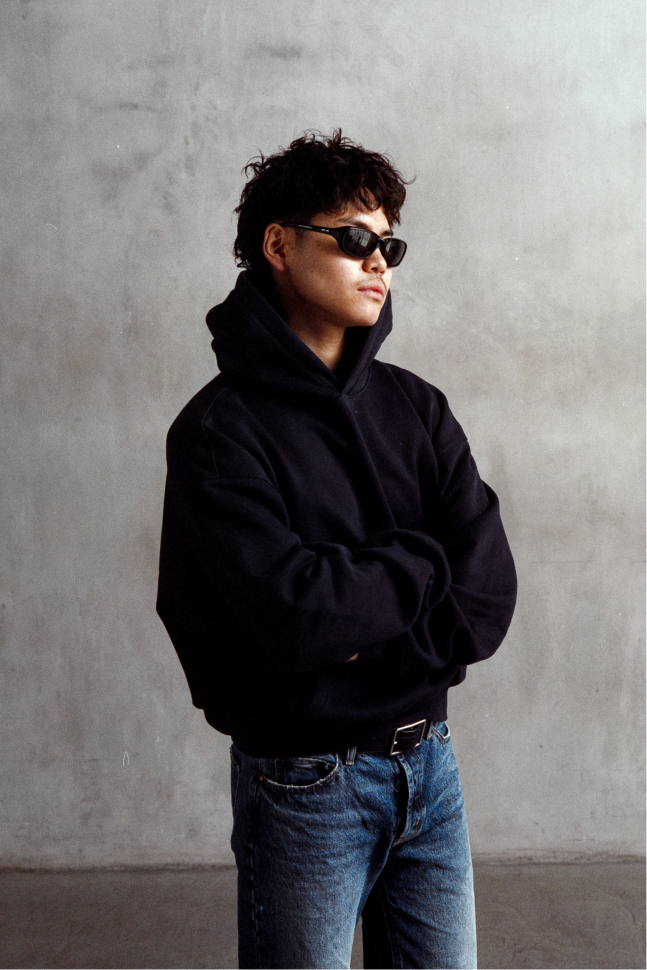Model wearing a black relaxed-fit hoodie with blue jeans and sunglasses, shown in a three-quarter view with folded arms.