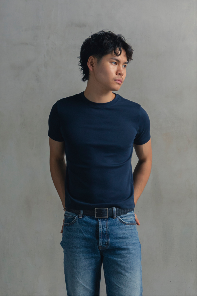 Model wearing a navy fitted crew neck T-shirt with blue jeans, looking to the side with hands behind the back.