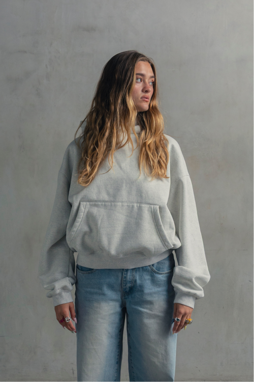 Female model wearing a light heather grey relaxed-fit hoodie with light blue jeans, shown from the front.