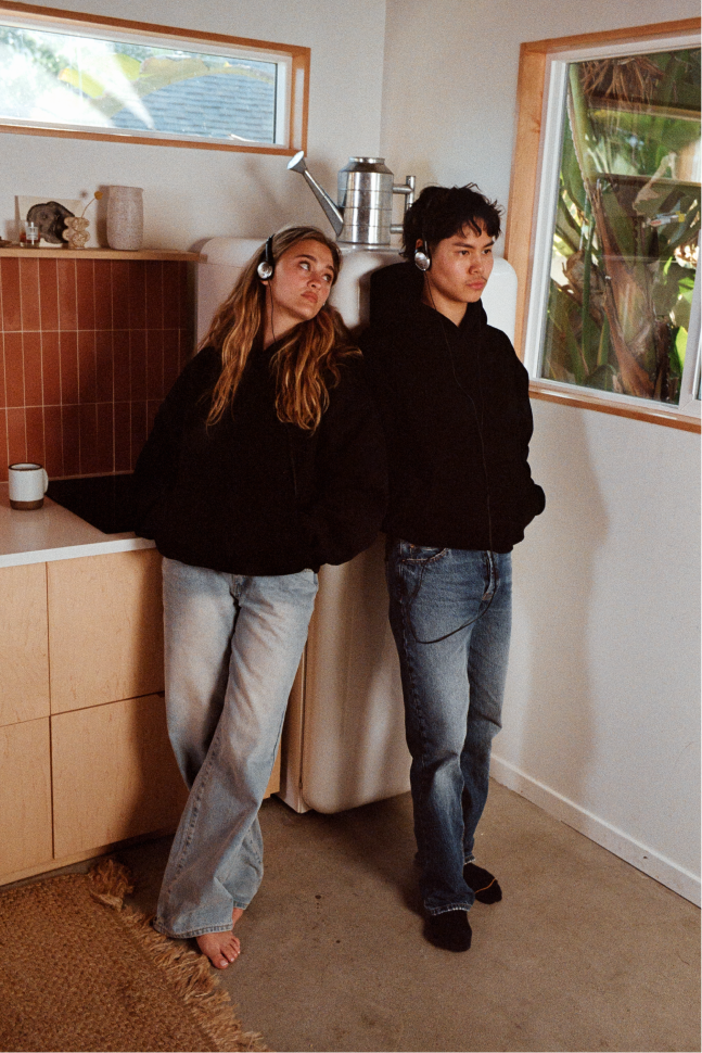 Two models standing barefoot in a kitchen corner, both wearing black hoodies, with one wearing light-wash flared jeans and the other medium-wash straight-leg jeans.