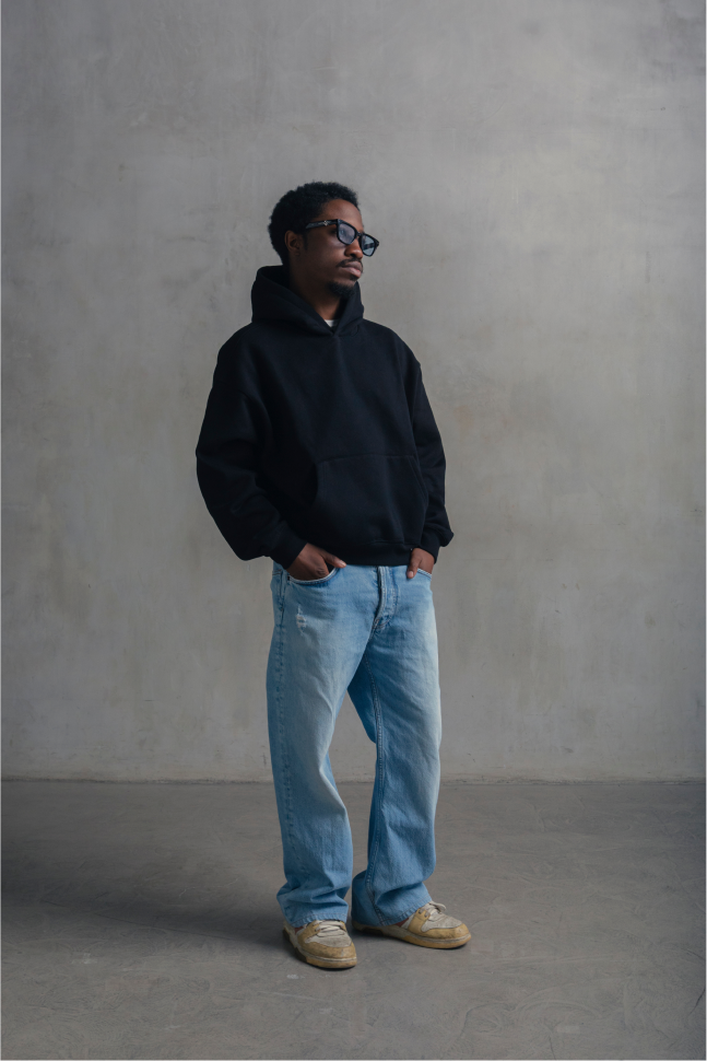 A man wearing a black hoodie, light blue jeans, and beige sneakers stands against a textured gray concrete wall.